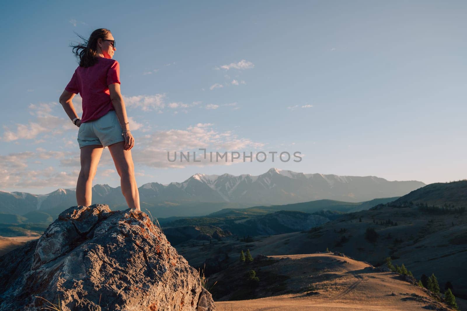A woman in casual clothing stands on a rocky peak, gazing at scenic mountains and a vast valley under golden sunlight.