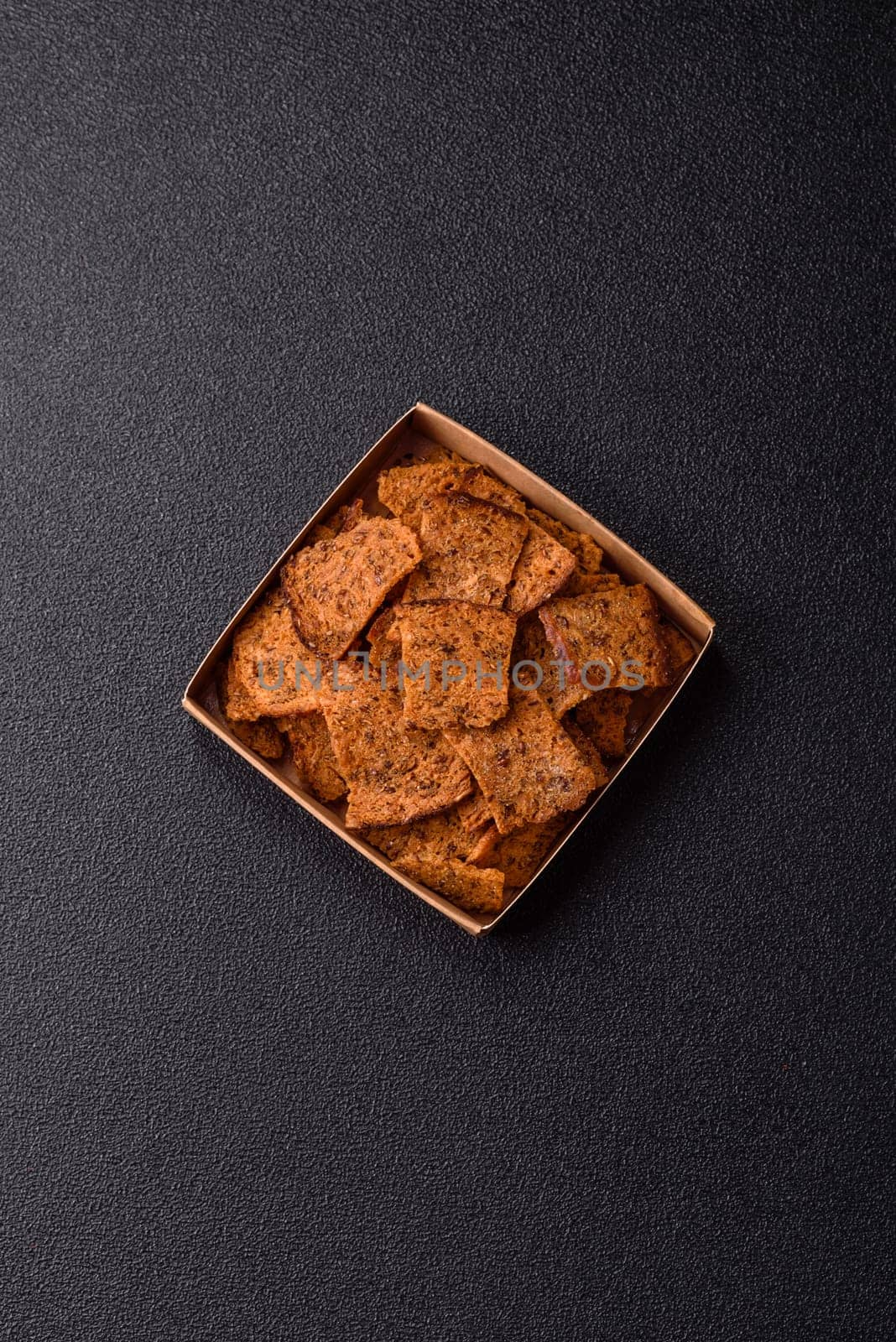 Crispy homemade brown bread chips with seeds and grains, salt, spices and herbs on a dark concrete background