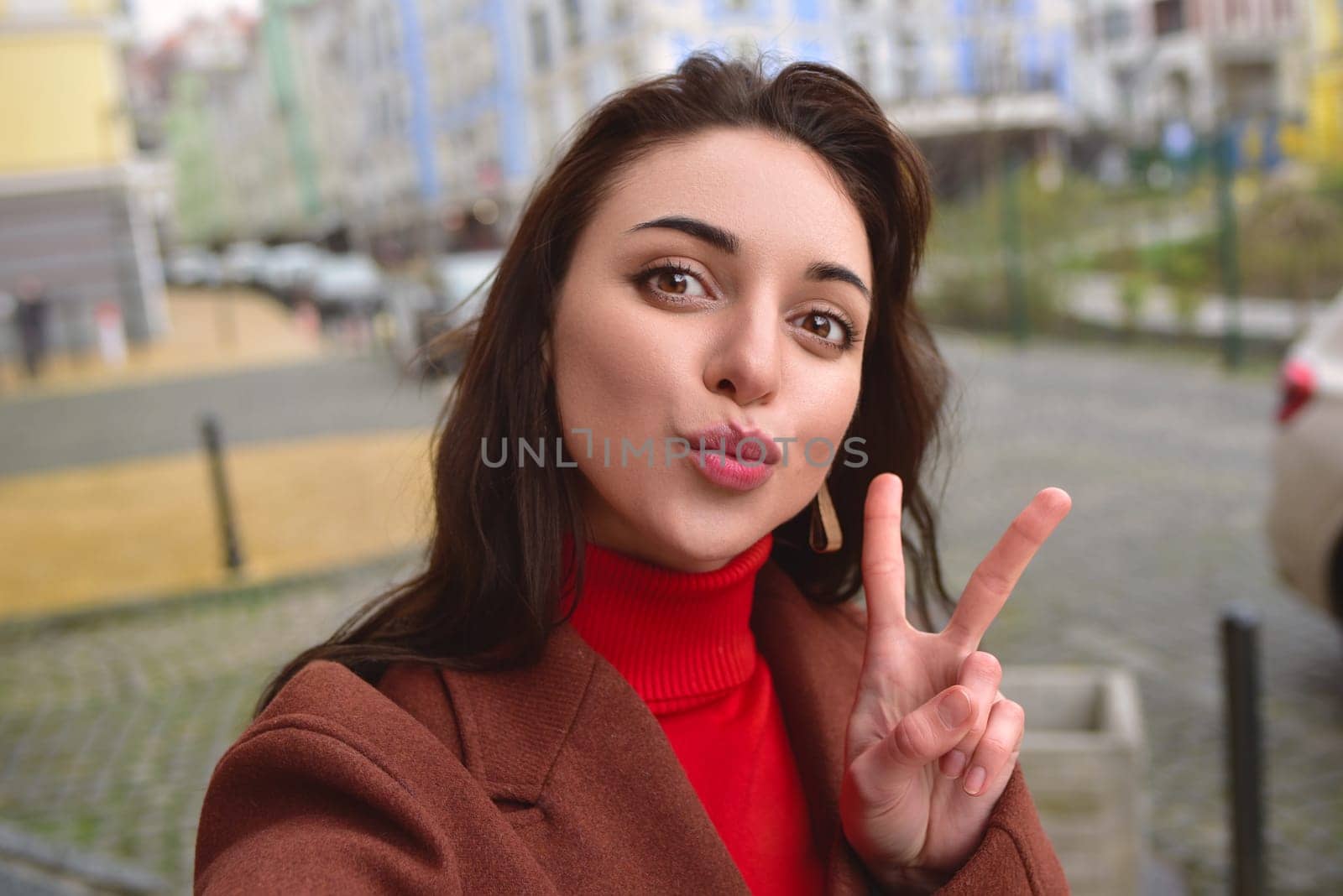 Charming lady in a brown autumn coat takes a selfie on the street with a peace sign by Nickstock
