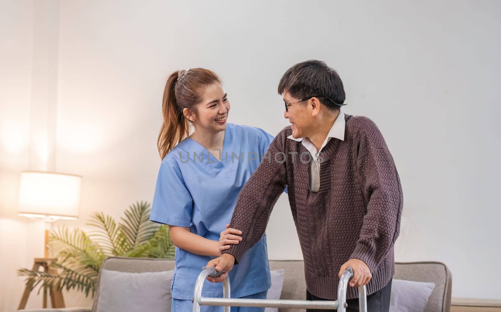 young asian female nurse care giver helping asian senior old man with mobility walker in living area of nursing home senior daycare center. Nurse take care elderly patient with cheerful concentrate by wichayada