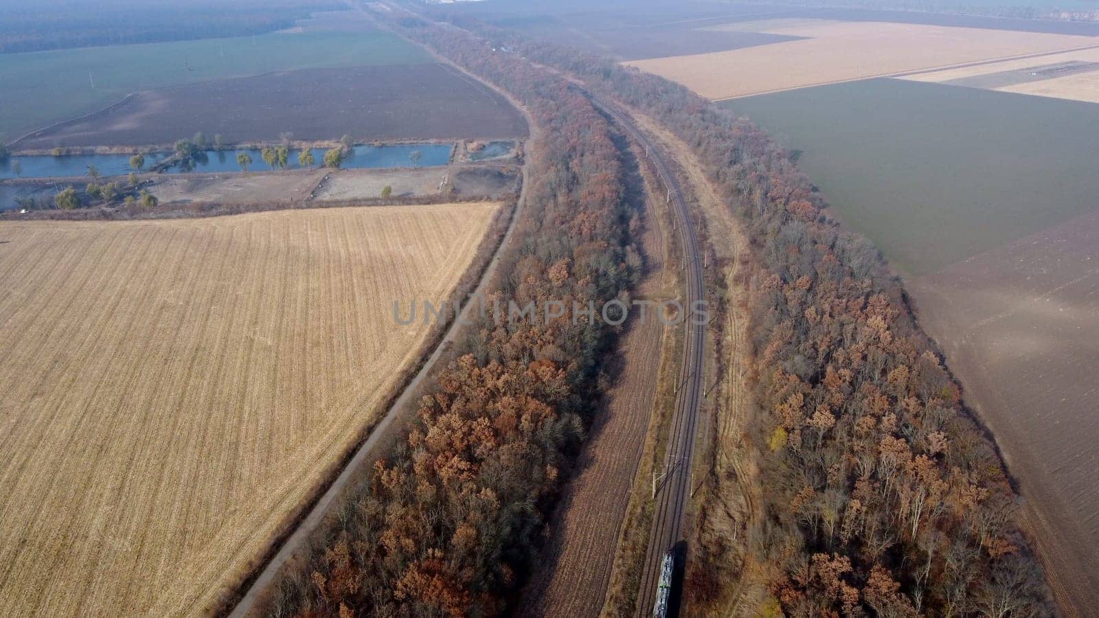 Panoramic View of Moving Freight Train Along Railway Tracks Among Trees Between Agricultural Fields on an Autumn Day. Landscape Freight Cars or Railway Wagon Rides on Railroad. Top View Rail freight