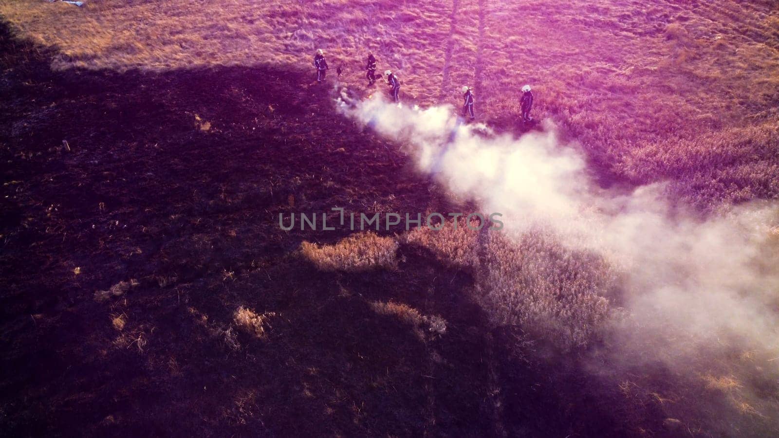 Group of firefighters puts out flame fire due to burning of dry grass in field on sunny day. View from above. Aerial drone view. Old dry grass burning on field. White smoke rising. Red pink sun glare