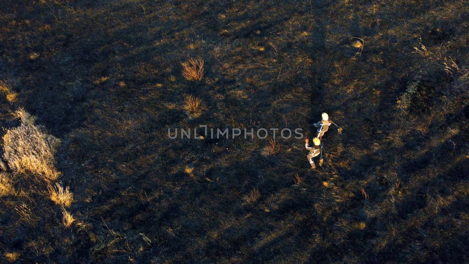 Two firefighters walk on black scorched earth after fire and burning dry grass in field. Two people in uniforms of firefighters walk on black earth and ash after extinguishing fire. Aerial drone view