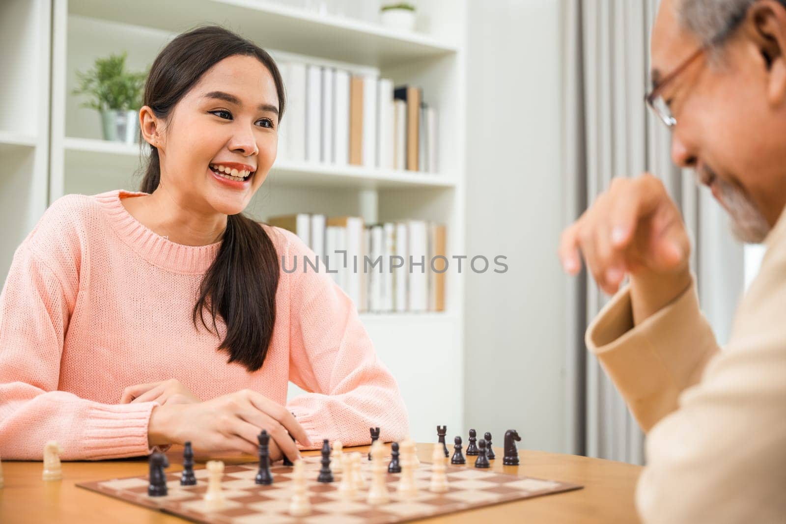 Beautiful young smile woman having fun sitting playing chess game with senior elderly at home by Sorapop