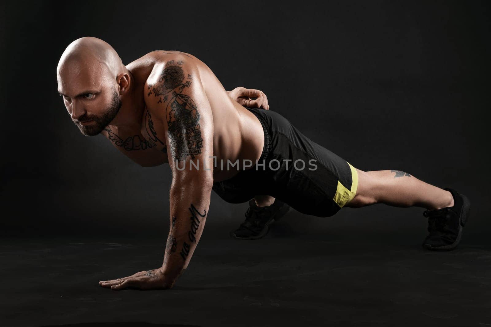 Masculine bald, bearded, tattooed man in black shorts and sneakers is pushing ups from the floor against a black background. Chic muscular body, fitness, gym, healthy lifestyle concept. Close-up portrait.