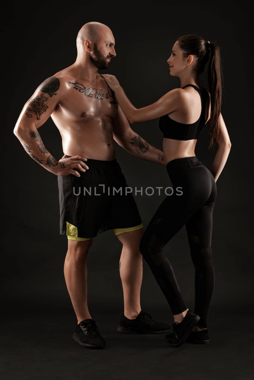 Strong bald, tattooed man in black shorts and sneakers with charming brunette woman in leggings and top are hugging standing sideways on black background and looking at each other. Fitness couple, chic muscular bodies, gym concept. The love story.