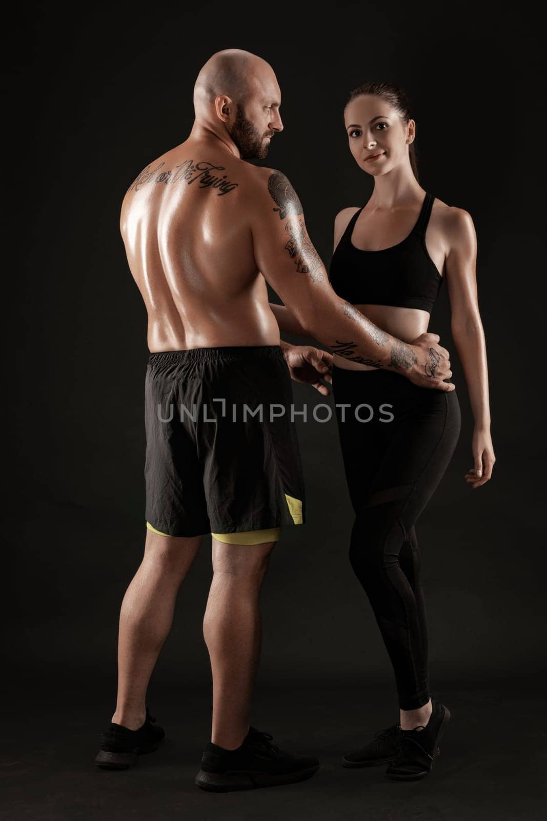 Strong bald, tattooed male in black shorts and sneakers, standing back, with charming brunette female in leggings and top are hugging on black background and looking at the camera. Fitness couple, chic muscular bodies, gym concept. The love story.
