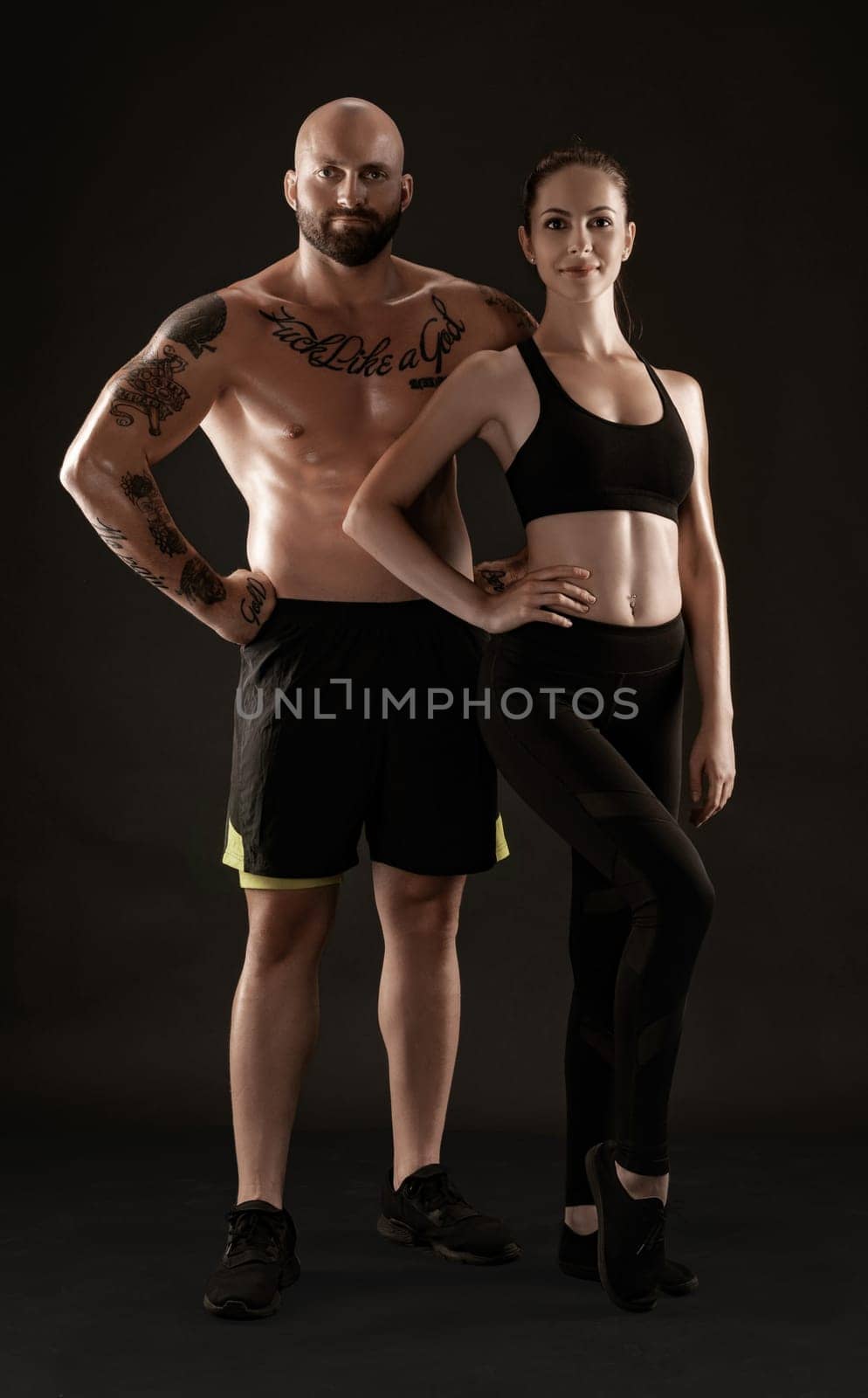 Strong bald, tattooed fellow in black shorts and sneakers with charming brunette lady in leggings and top are posing on black background and looking at the camera. Fitness couple, chic muscular bodies, gym concept. The love story.