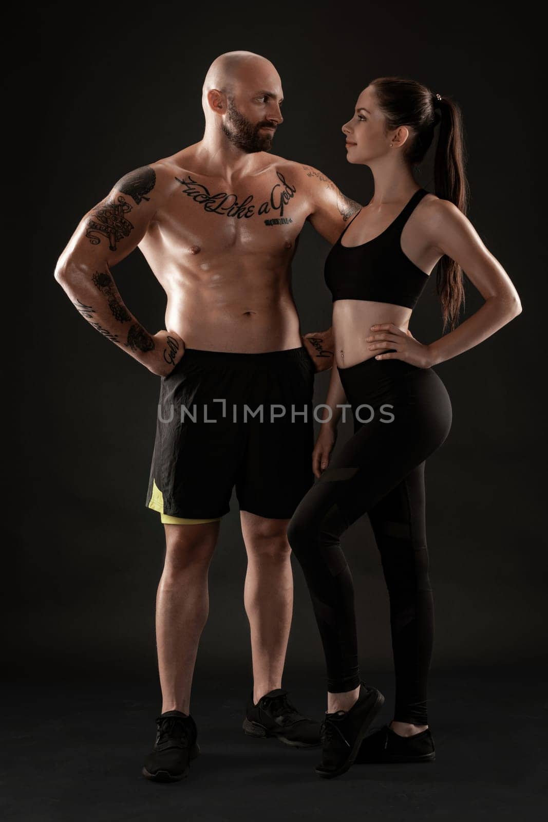 Athletic man in shorts and sneakers with brunette woman in leggings and top posing on black background. Fitness couple, gym concept. by nazarovsergey