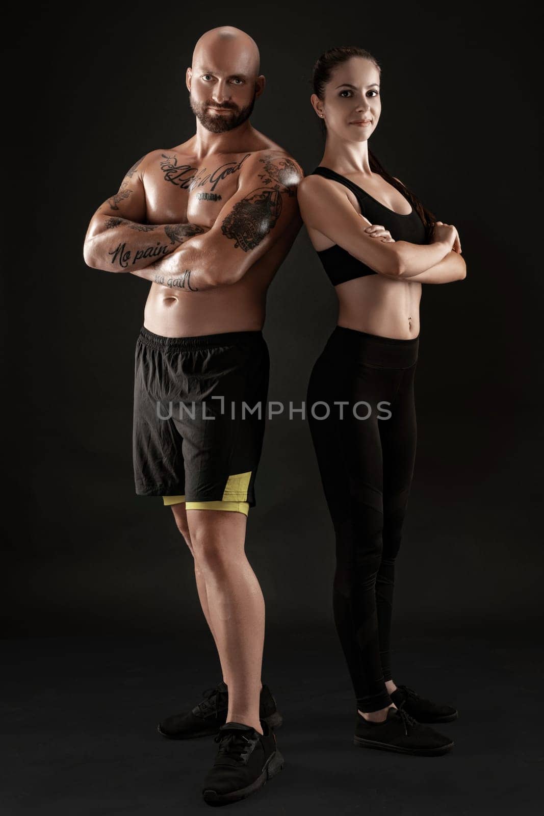 Handsome bald, tattooed male in black shorts and sneakers with gorgeous brunette female in leggings and top are posing with crossed hands on black background and looking at the camera. Fitness couple, chic muscular bodies, gym concept. The love story.