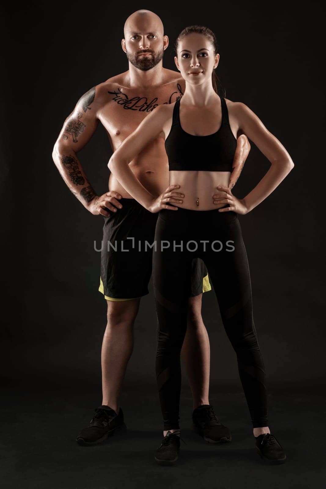 Handsome bald, tattooed fellow in black shorts and sneakers with gorgeous brunette lady in leggings and top are posing on black background and looking at the camera. Fitness couple, chic muscular bodies, gym concept. The love story.