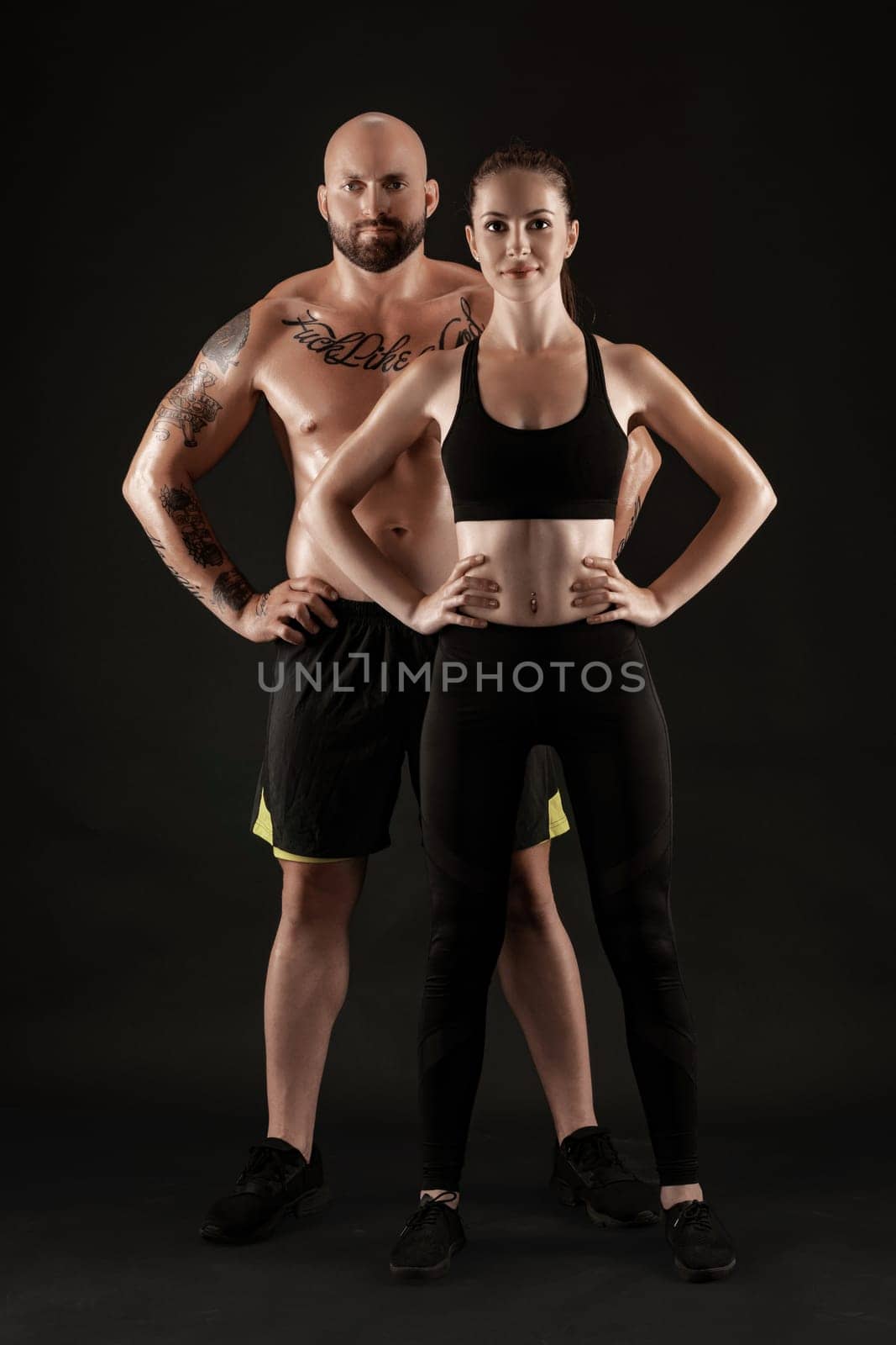 Handsome bald, tattooed man in black shorts and sneakers with gorgeous brunette maiden in leggings and top are posing on black background and looking at the camera. Fitness couple, chic muscular bodies, gym concept. The love story.