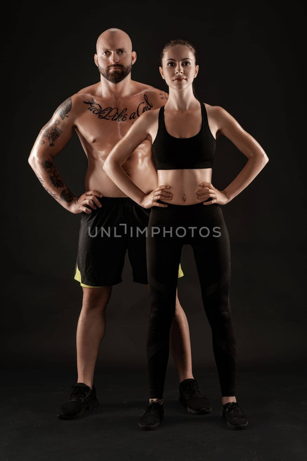 Good-looking bald, tattooed man in black shorts and sneakers with attractive brunette woman in leggings and top are posing on black background and looking at the camera. Fitness couple, chic muscular bodies, gym concept. The love story.