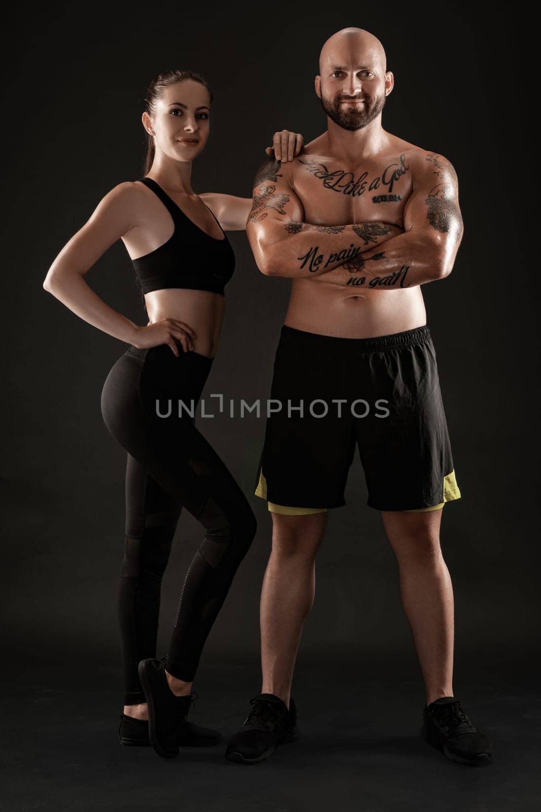 Good-looking bald, tattooed guy in black shorts and sneakers with attractive brunette girl in leggings and top are posing on black background and looking at the camera. Fitness couple, chic muscular bodies, gym concept. The love story.