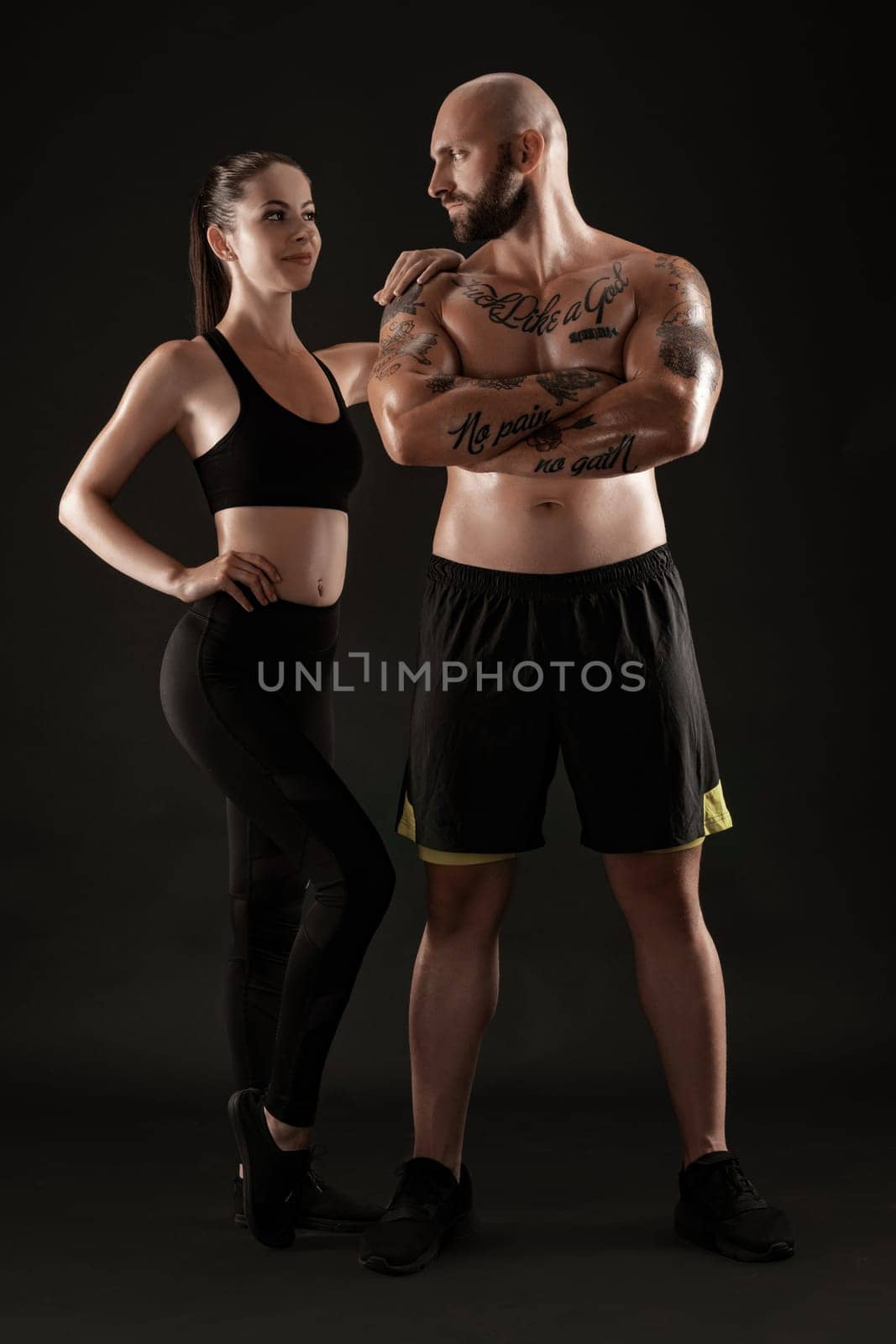 Good-looking bald, tattooed man in black shorts and sneakers with attractive brunette maiden in leggings and top are posing on black background and looking at each other. Fitness couple, chic muscular bodies, gym concept. The love story.