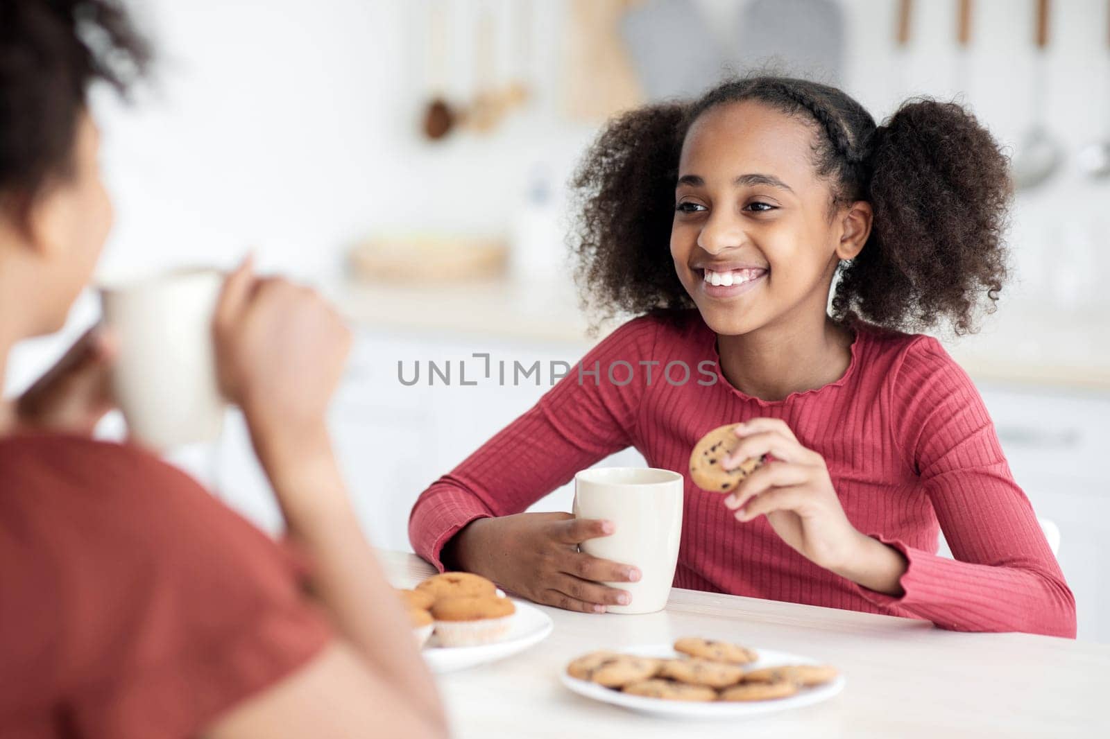 Cute african american teen girl drinking tea with her mom, sitting at kitchen table, eating biscuits, cookies, muffins, having pleasure conversation and smiling, selective focus on black kid