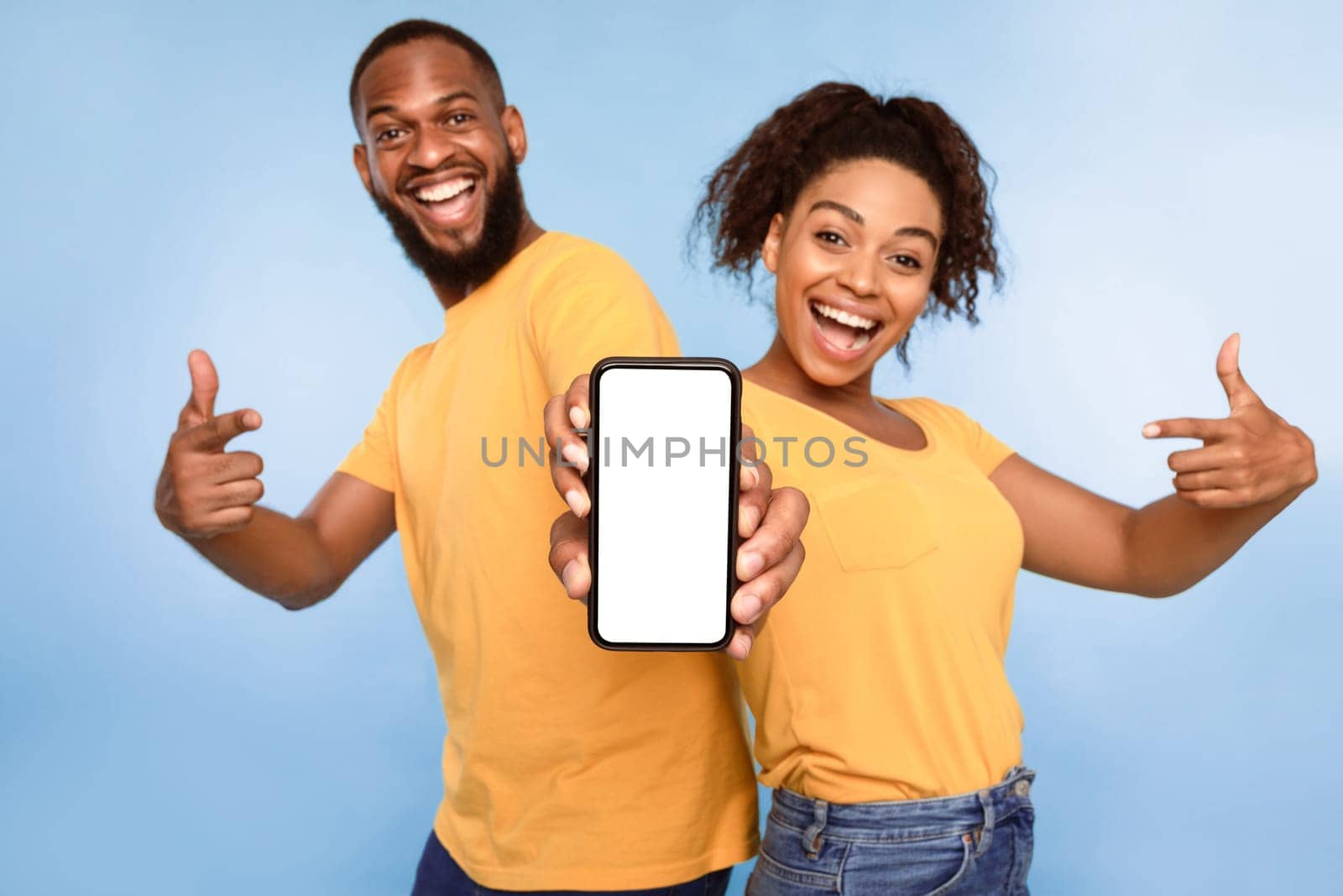 Mobile application ad. Excited African American couple pointing at cellphone with blank screen, recommending new app on blue studio background, selective focus. Mockup for website
