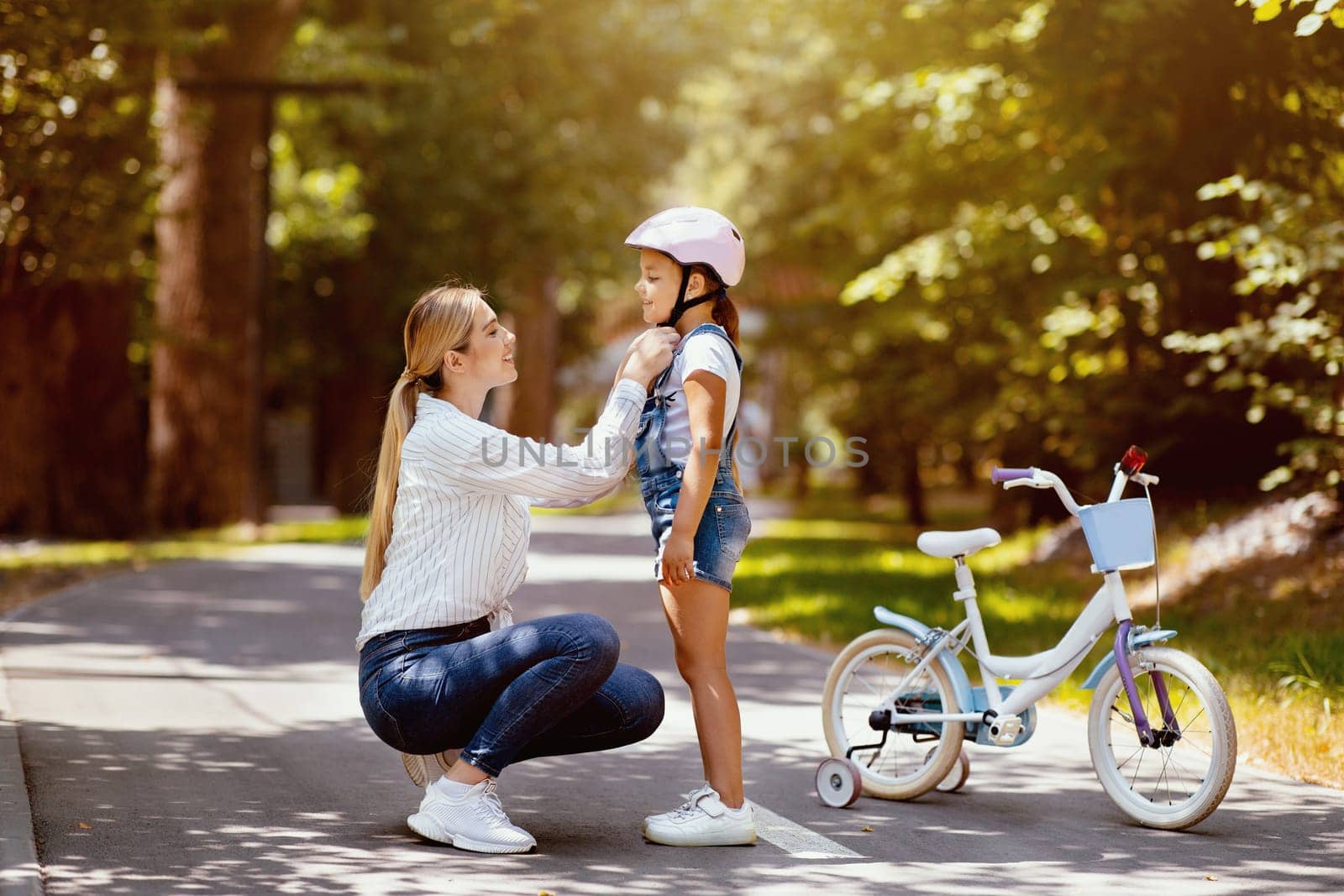 Mother Putting Helmet On Daughter Teaching Her To Ride A Bicycle In Park On Weekend. Free Space For Text