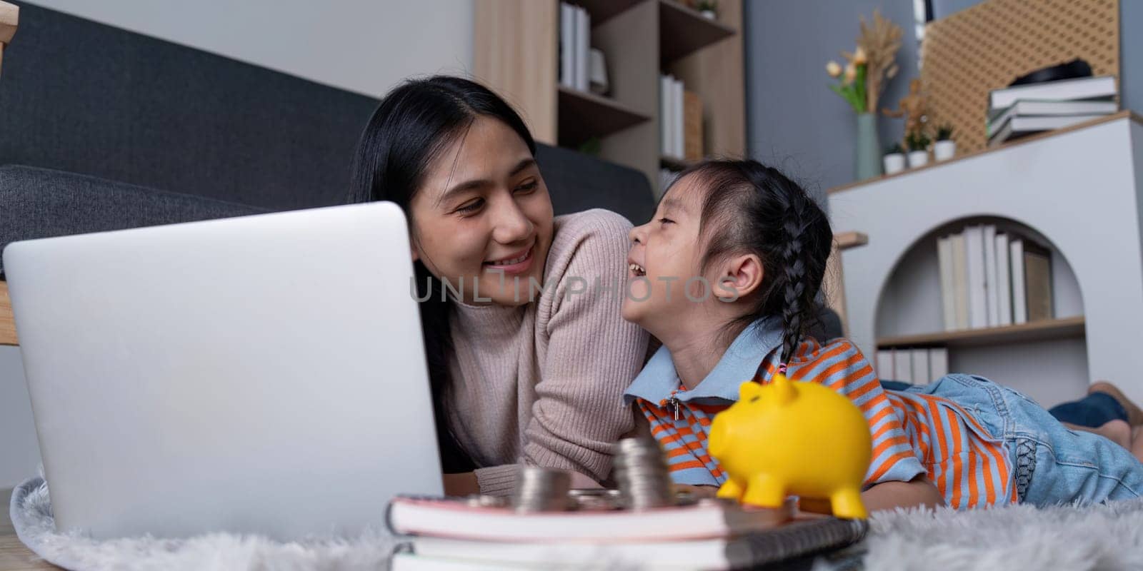 A diverse mother and her daughter share a joyful moment as they engage with a yellow piggy bank and laptop, promoting financial education and family bonding at home.
