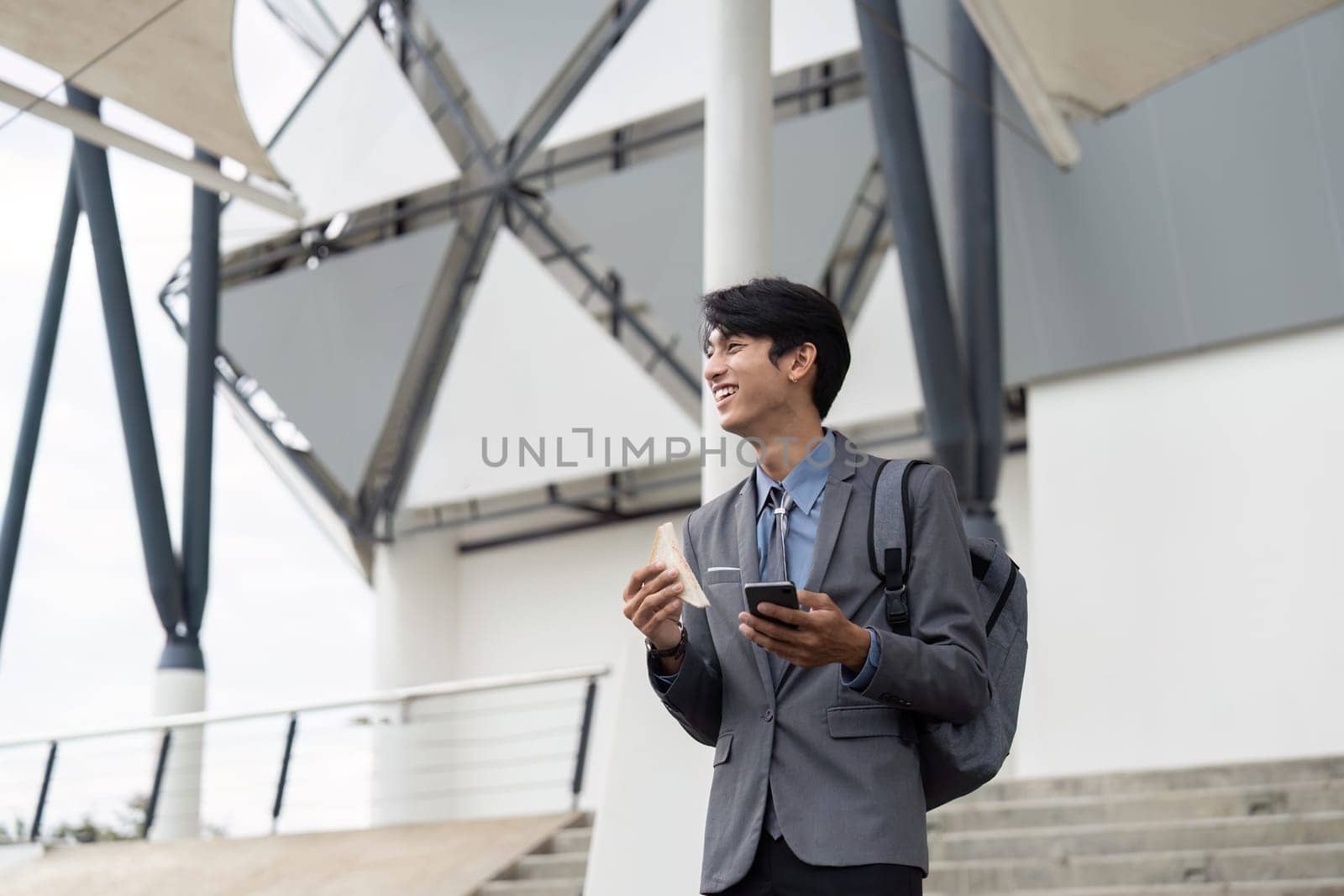 A young Asian businessman in a suit, smiling as he enjoys a snack and checks his smartphone, embodying a cheerful and balanced work life in a contemporary outdoor environment.