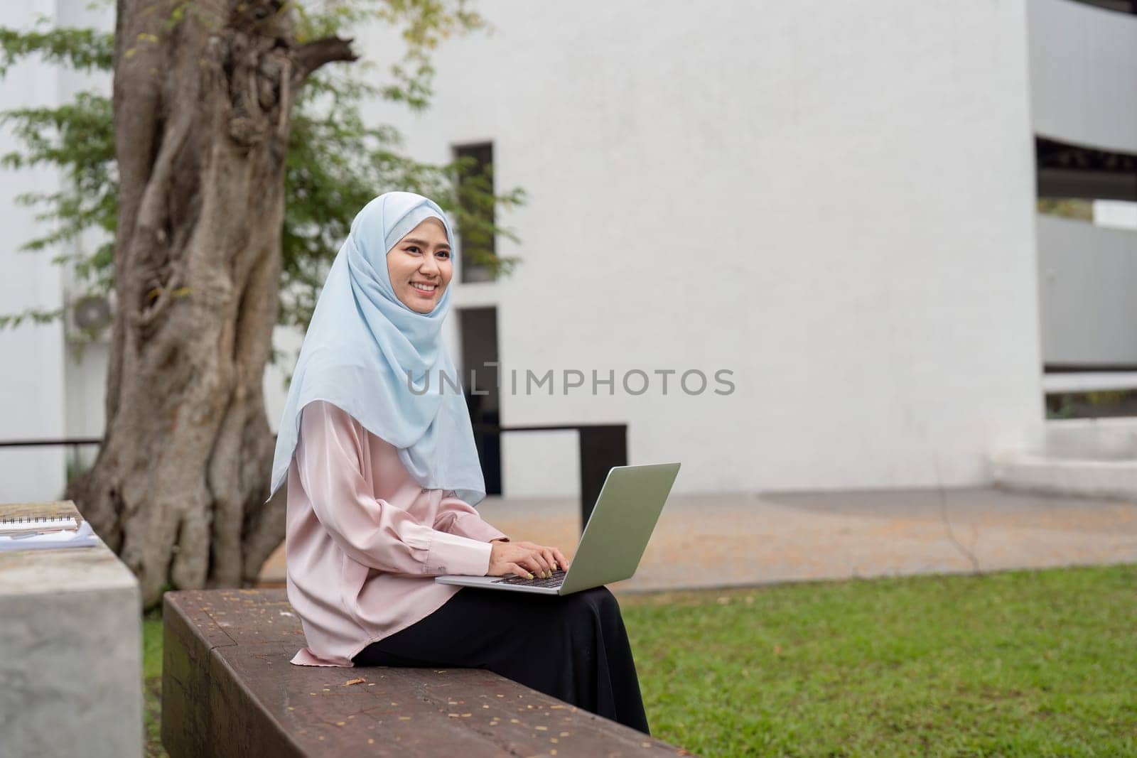 A cheerful Muslim businesswoman in a hijab, happily working on her laptop while seated on a bench in a green outdoor space.