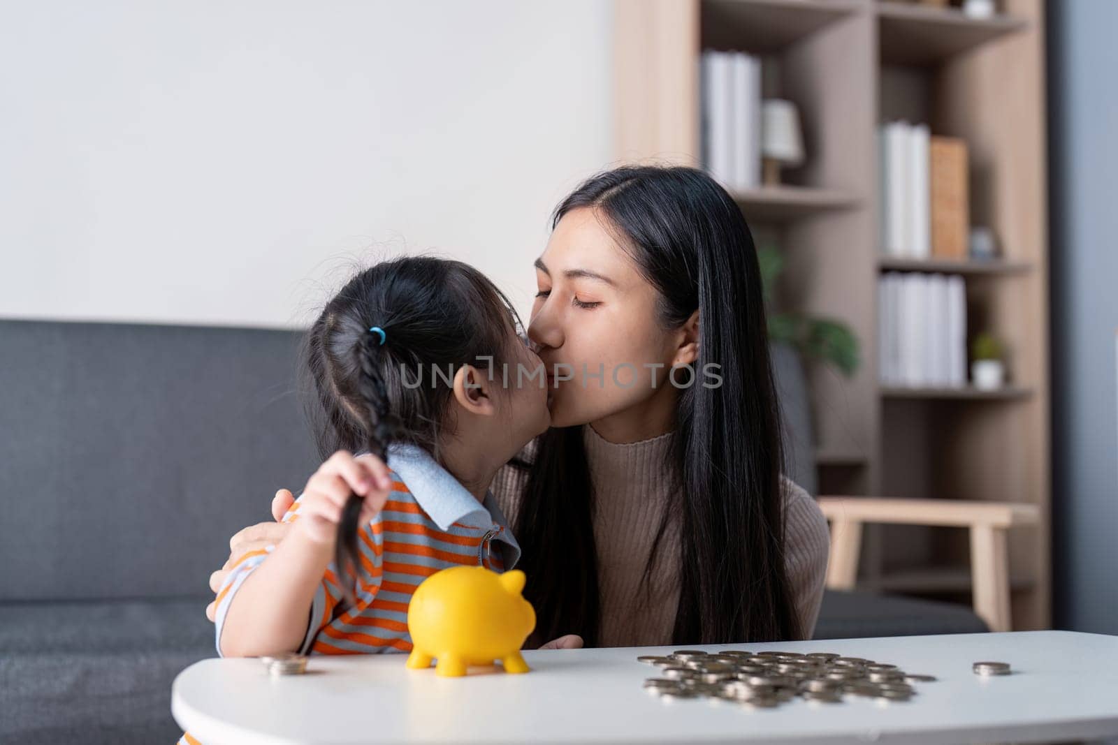 A diverse mother and her daughter share a sweet kiss as they engage in saving money together with a yellow piggy bank and coins, enhancing their family bond.