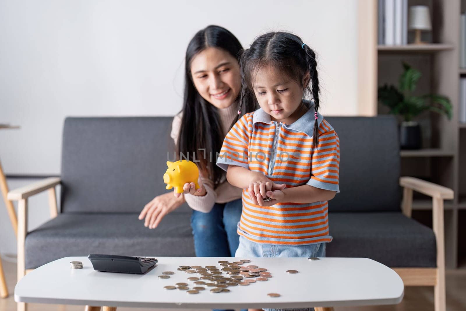 A mother and her daughter joyfully count coins on a table, engaging in a fun lesson about saving money in their stylish living room.