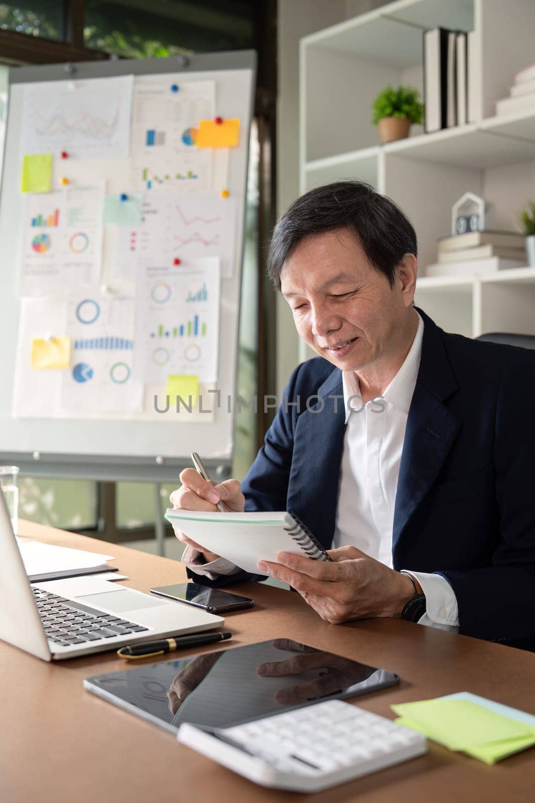 An elderly Asian businessman happily taking notes in his notebook, engaged in a meeting with visual data in the background.