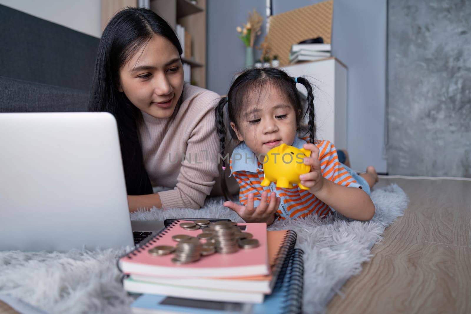 A diverse mother and her daughter examine a yellow piggy bank and coins together, enhancing their understanding of saving money while using a laptop at home.