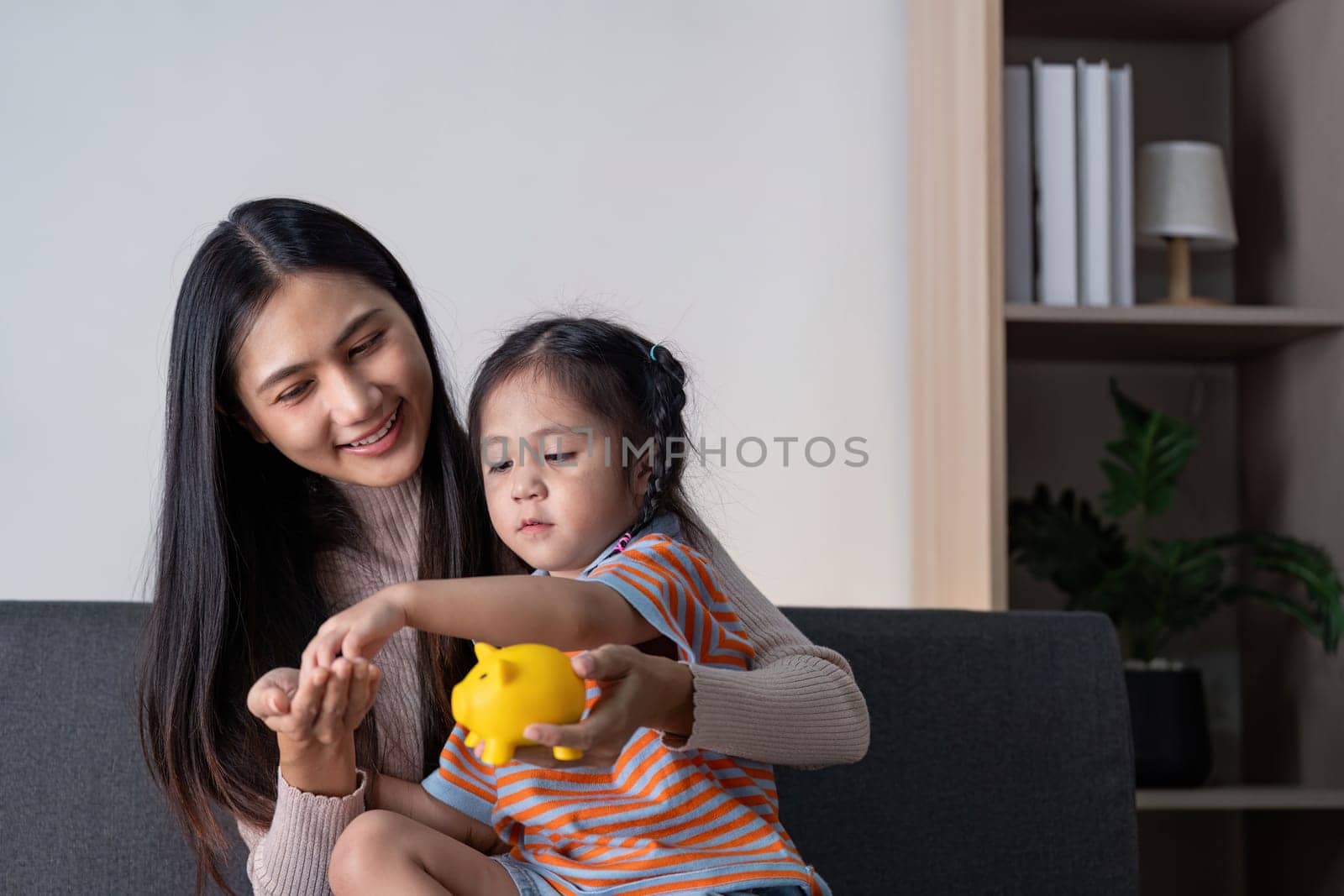 A diverse mother and her daughter engage in a fun activity, using a yellow piggy bank to learn about saving money together at home.