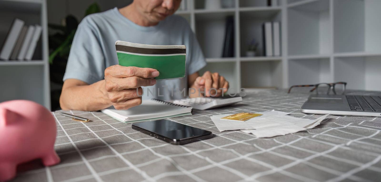 Elderly man thoughtfully analyzing his savings, holding a wallet while using a calculator and surrounded by essential documents in his home office.