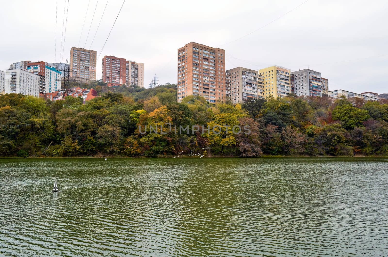 Dull and bleak view of old multi-story houses, glum sky, autumn forest and the lake