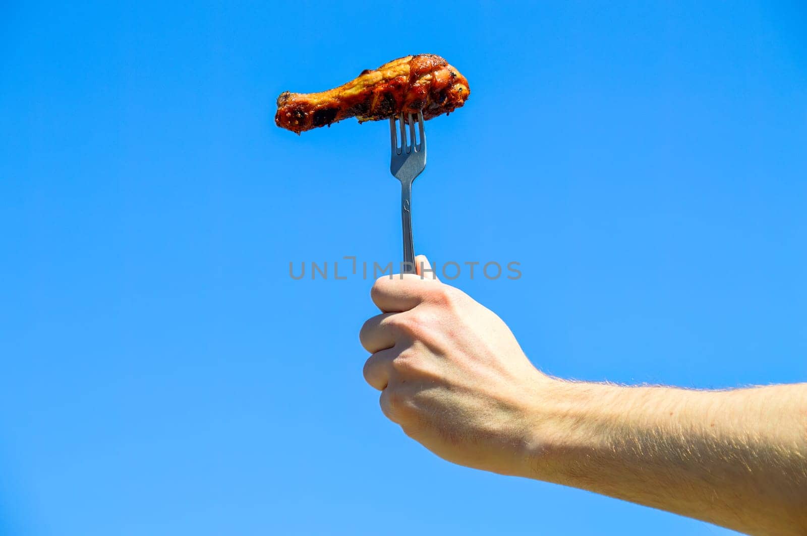 Hand holding fork with grilled appetizing chicken leg against the blue sky. Outdoor barbecue