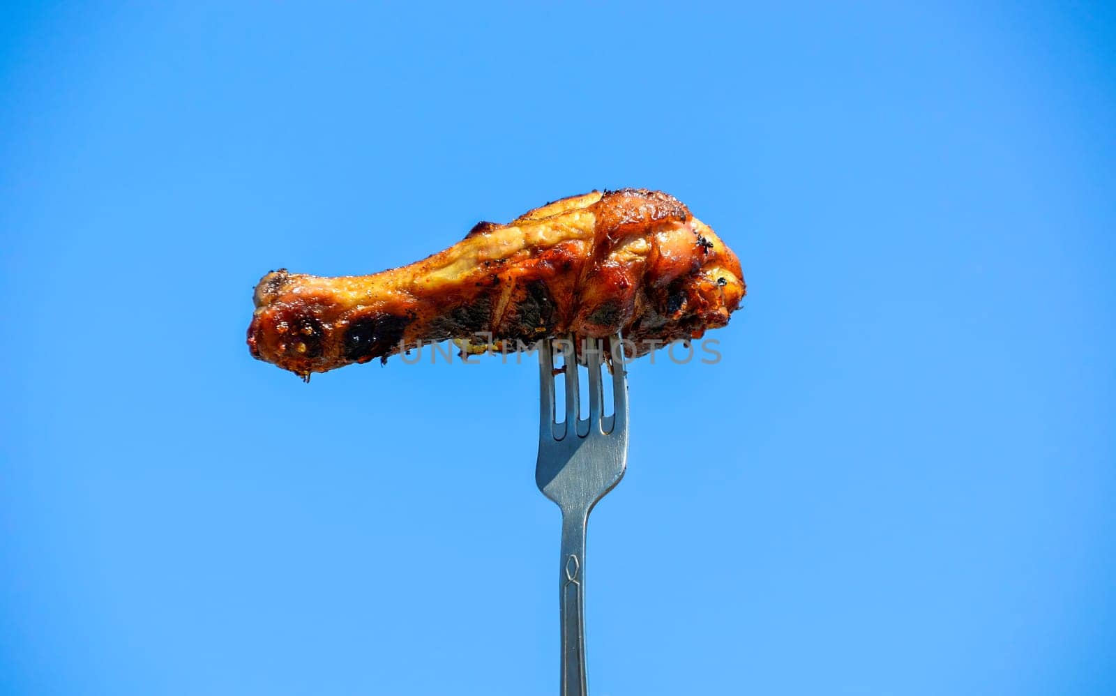 Grilled appetizing chicken leg on the fork against the blue sky. Outdoor barbecue