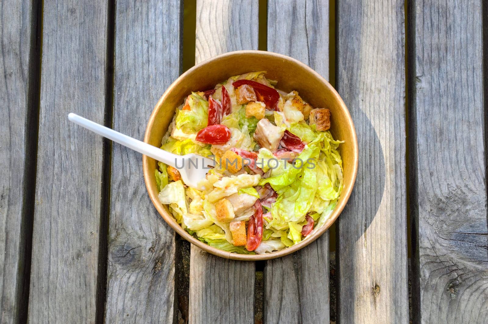 Paper bowl of fresh caesar salad with a fork on a wooden table. takeaway food