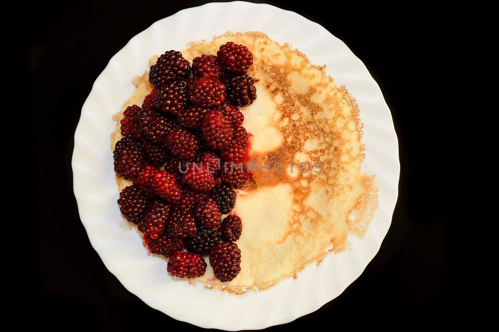 Ripe blackberries on ruddy appetizing pancake on white glass plate and black table