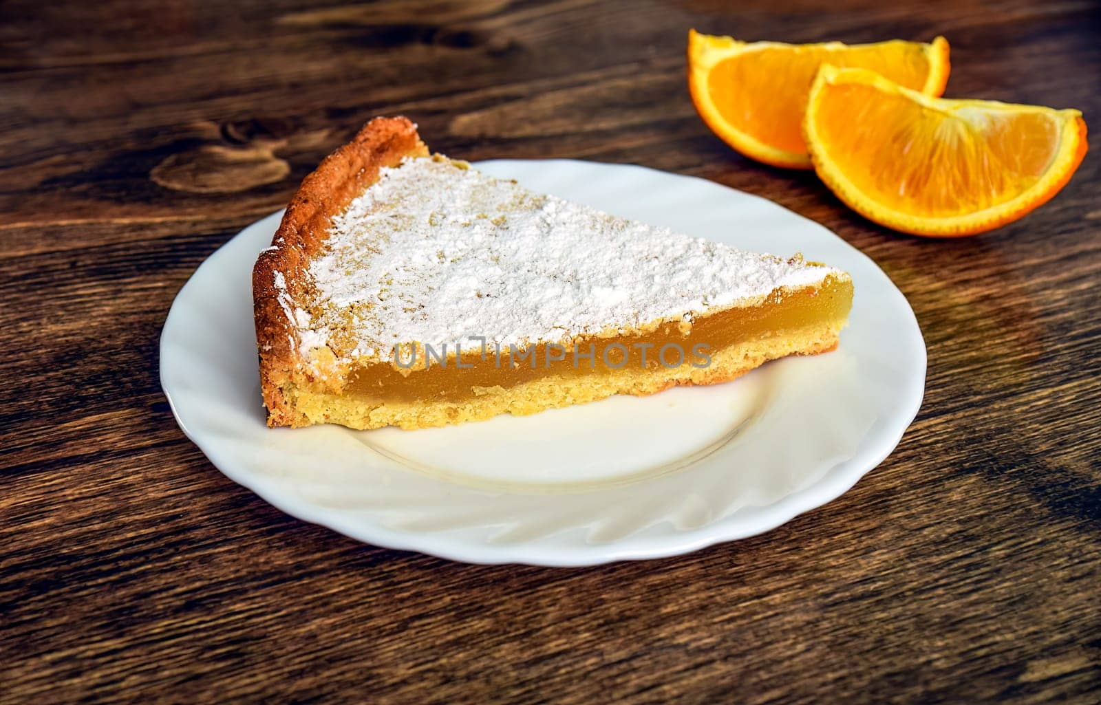 Piece of homemade pie with lemon and orange filling, covered with powdered sugar on a wooden table near orange fruit and cup of tea