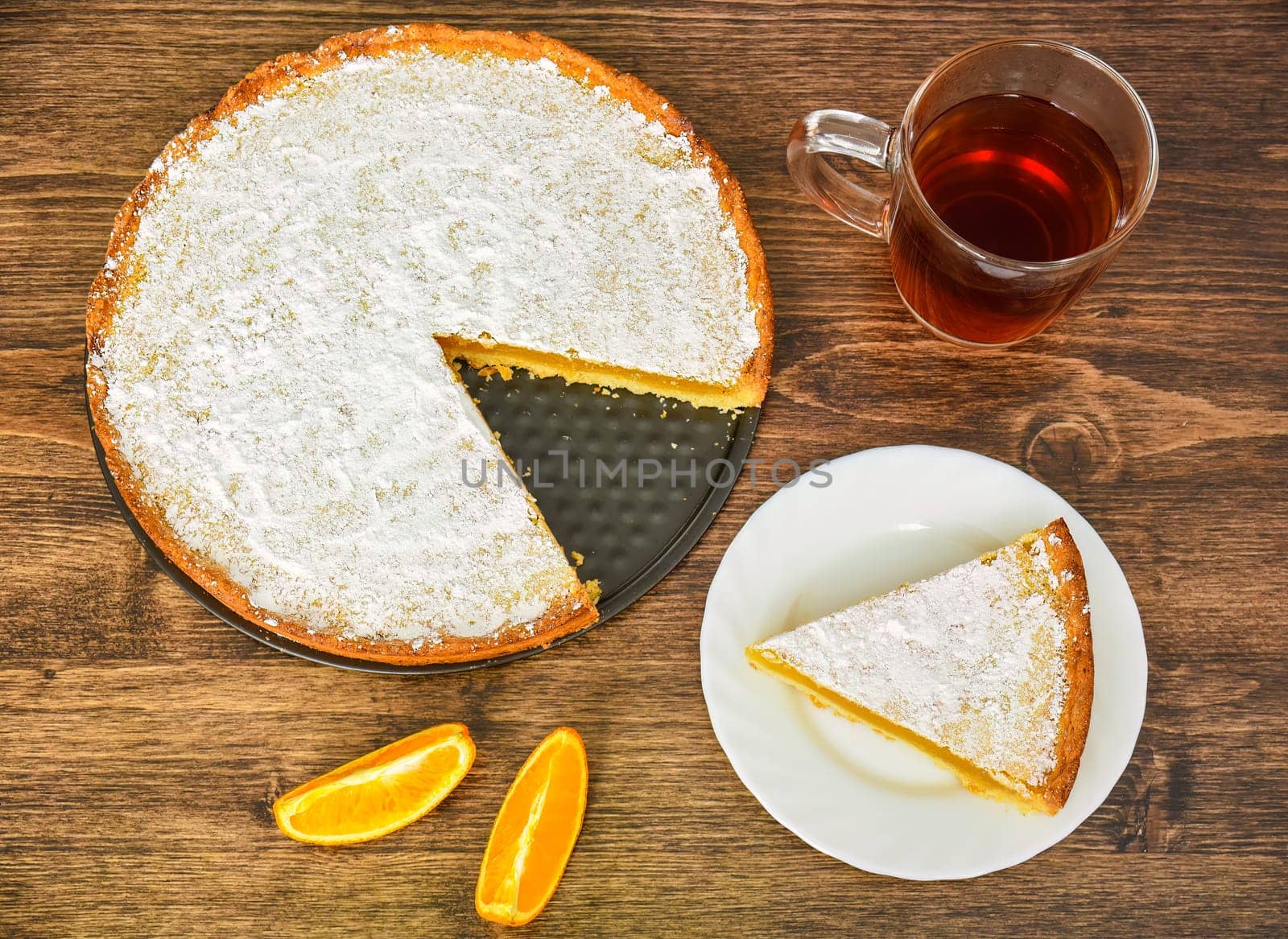 Piece of homemade pie with lemon and orange filling, covered with powdered sugar on a wooden table near orange fruit and cup of tea