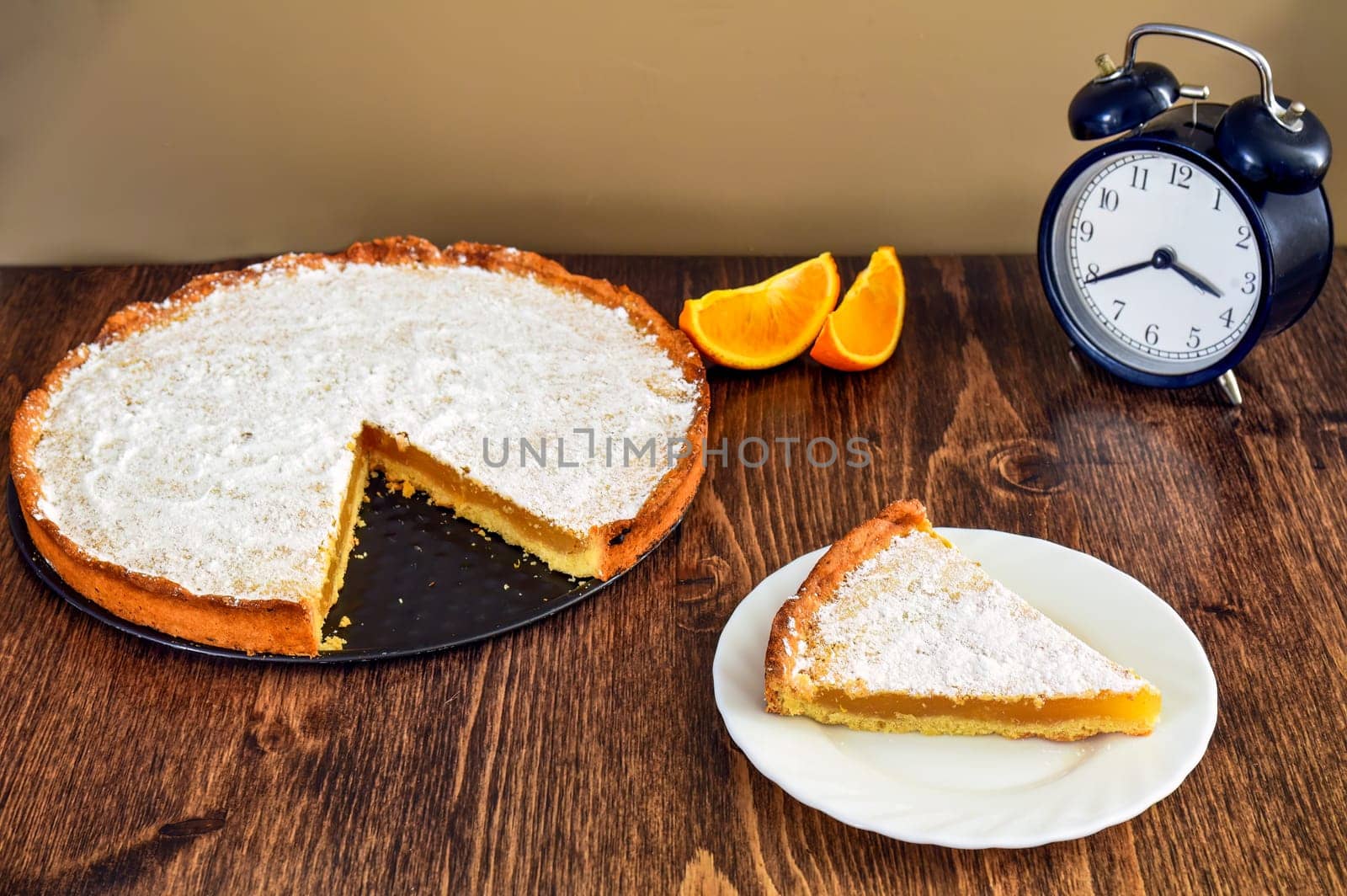 Piece of homemade pie with lemon and orange filling, covered with powdered sugar on a wooden table near orange fruit and alarm clock