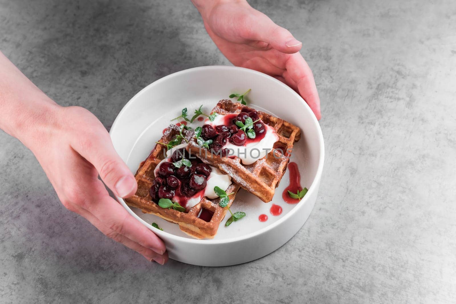Waiter serving white plate with sweet belgian waffles with cream and cherry jam on a gray table in a cafe