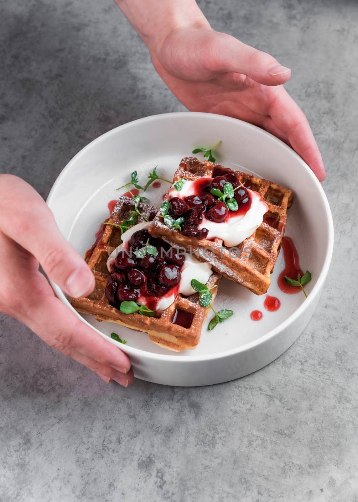 Waiter serving white plate with sweet belgian waffles with cream and cherry jam on a gray table in a cafe