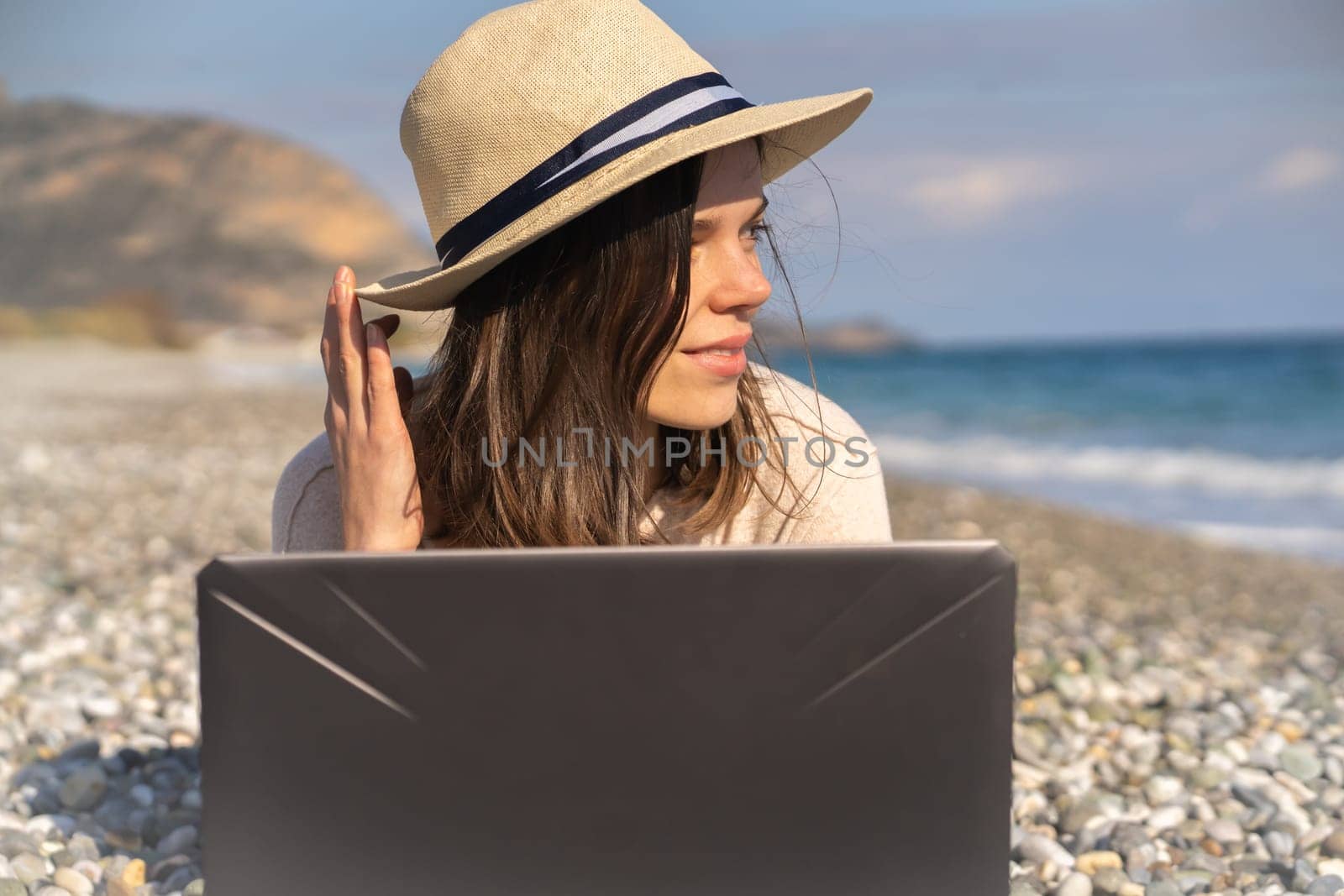 A young girl in a light hat and casual sweater lies on the beach by the sea with a laptop on a sunny day, works, studies, buys tickets during trip, a woman rests on vacation and types on the keyboard.