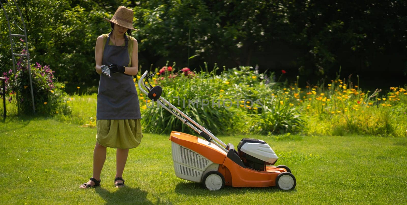 A young girl in a straw hat is mowing a lawn in the backyard with an orange lawn mower. A woman gardener is trimming grass with the grass cutter. A lawnmower is cutting a lawn on a summer sunny day.