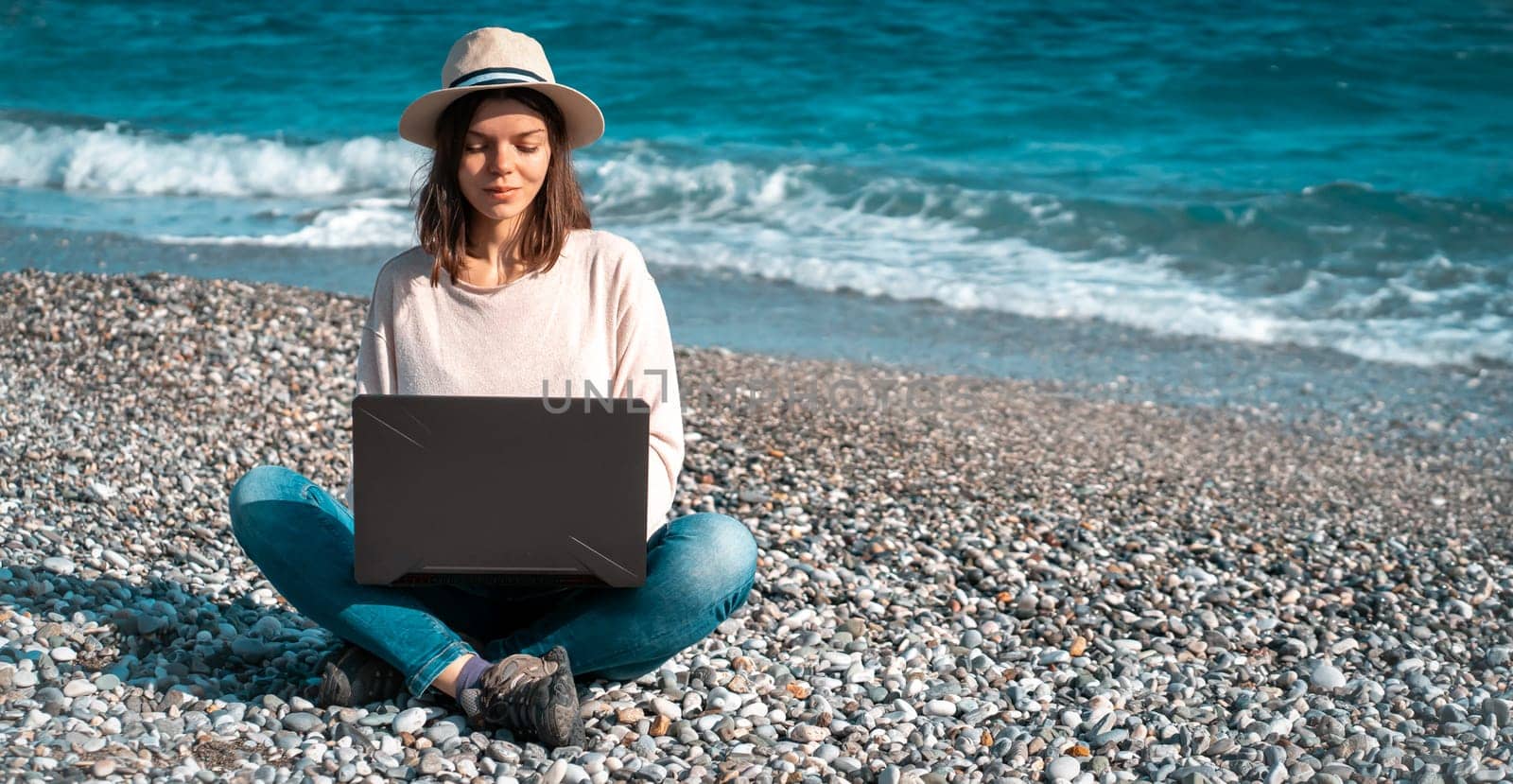 A young girl in a light hat and casual sweater lies on the beach by the sea with a laptop on a sunny day, works, studies, buys tickets during trip, a woman rests on vacation and types on the keyboard.
