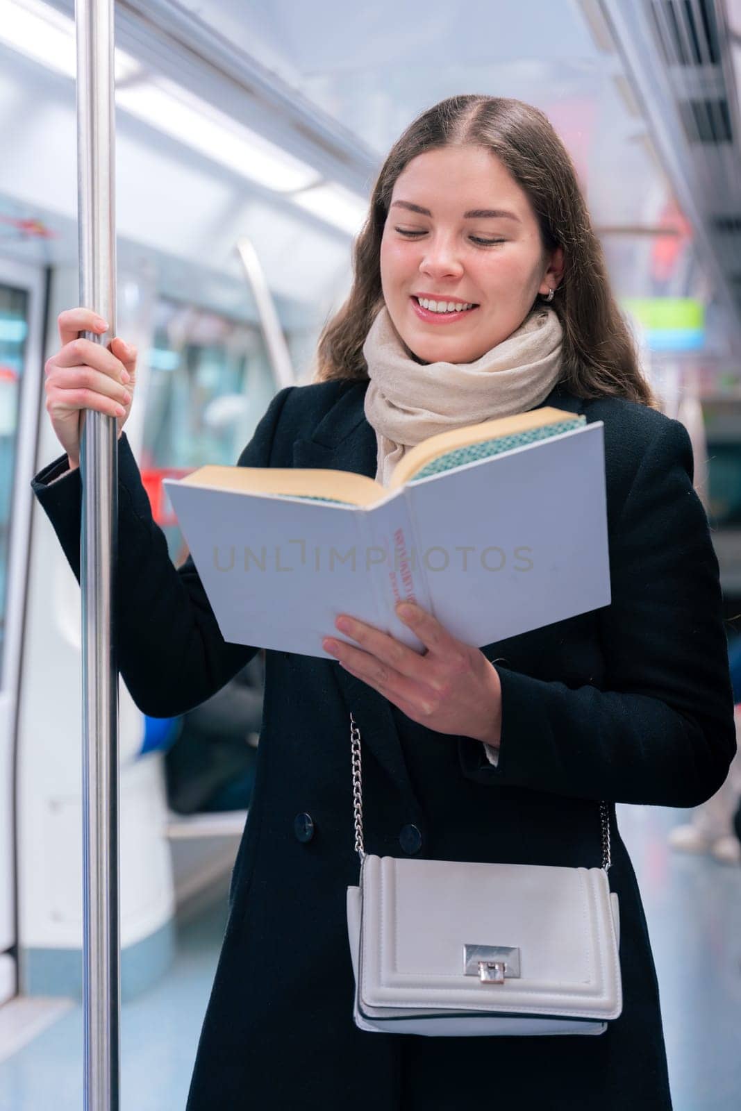 Focused young woman in a black coat and beige scarf standing inside a modern subway train, reading a book while holding onto a metal pole. She enjoys a quiet moment during her commute.