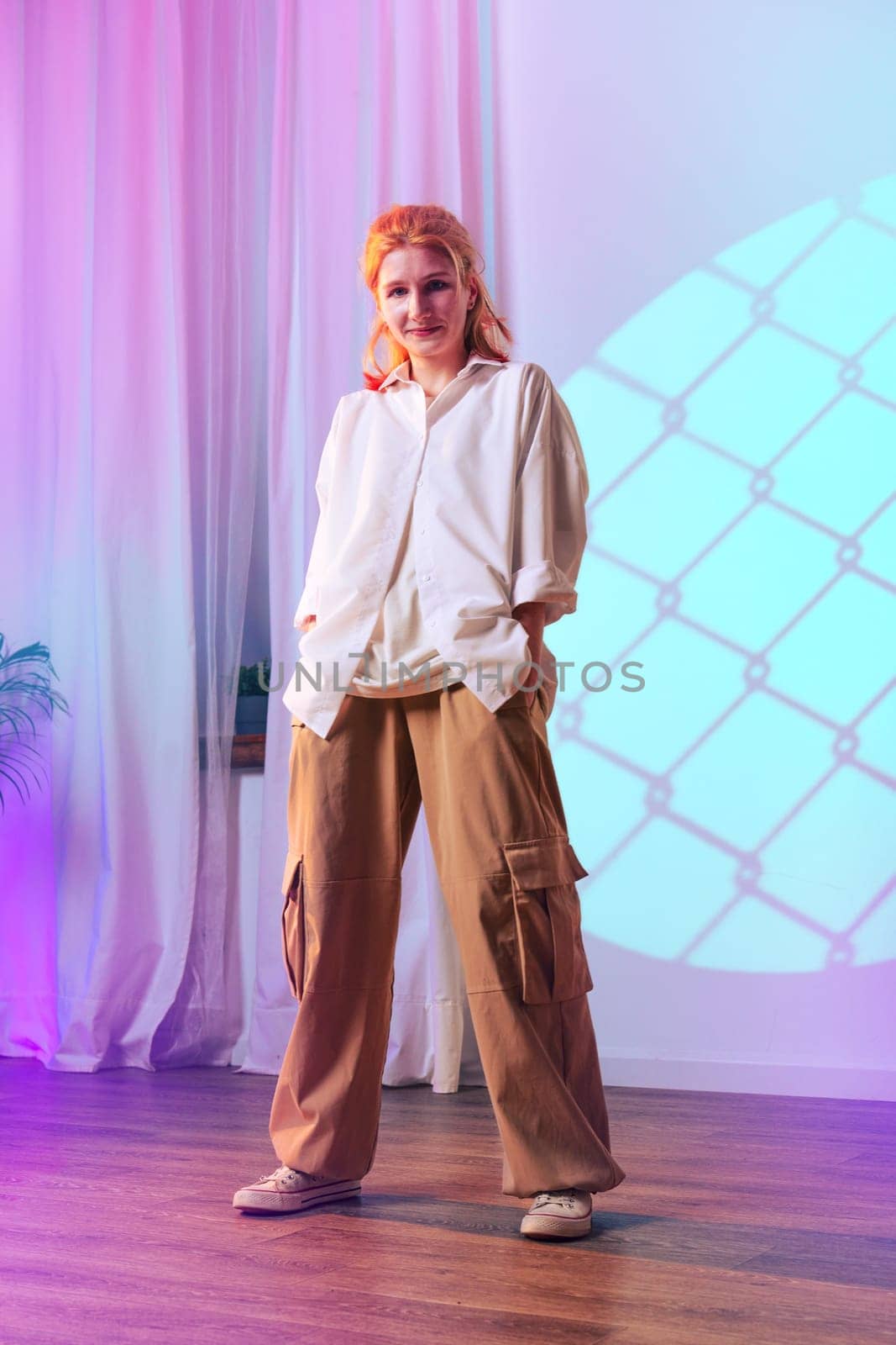 A woman stands confidently, wearing a loose white shirt and tan cargo pants. She poses in an indoor space highlighted with pastel lighting and decorative curtains, creating a trendy atmosphere.