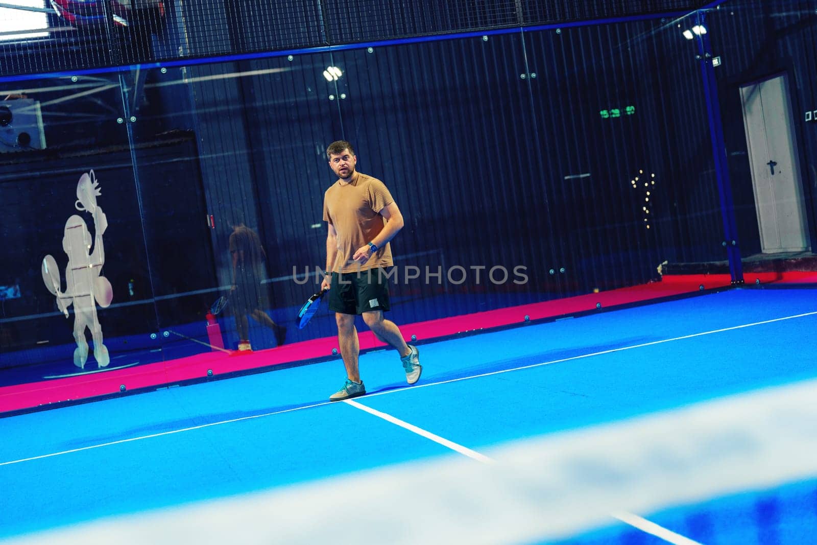 A young man engages in a friendly game of padel tennis on a striking blue court in an indoor sports facility, showcasing his skills and enthusiasm for the sport.