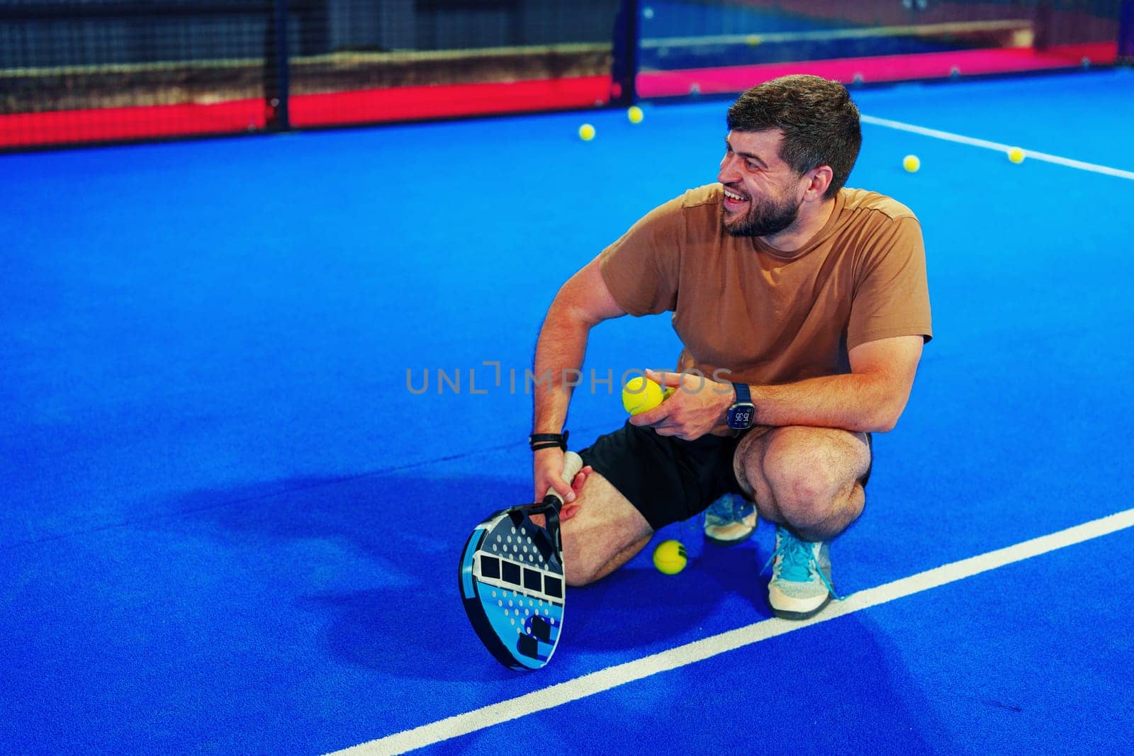 A man crouches on a bright blue court, smiling as he holds a paddle and tennis balls. He seems to be enjoying a fun practice session, surrounded by scattered yellow balls.