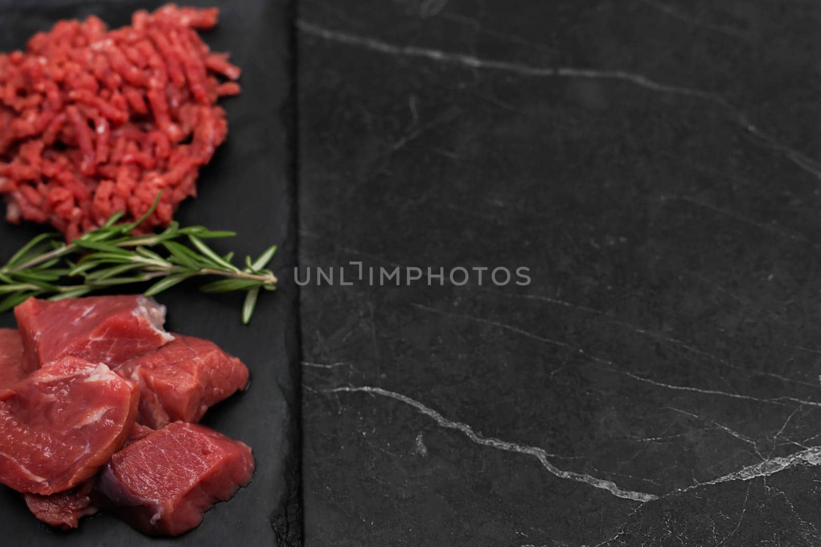 Top left view of chunks of beef lying on a black cutting board, separated by sprigs of rosemary with red minced meat on a dark background with space for text. The concept of cooking, shredding meat, diet, natural products. High quality photo