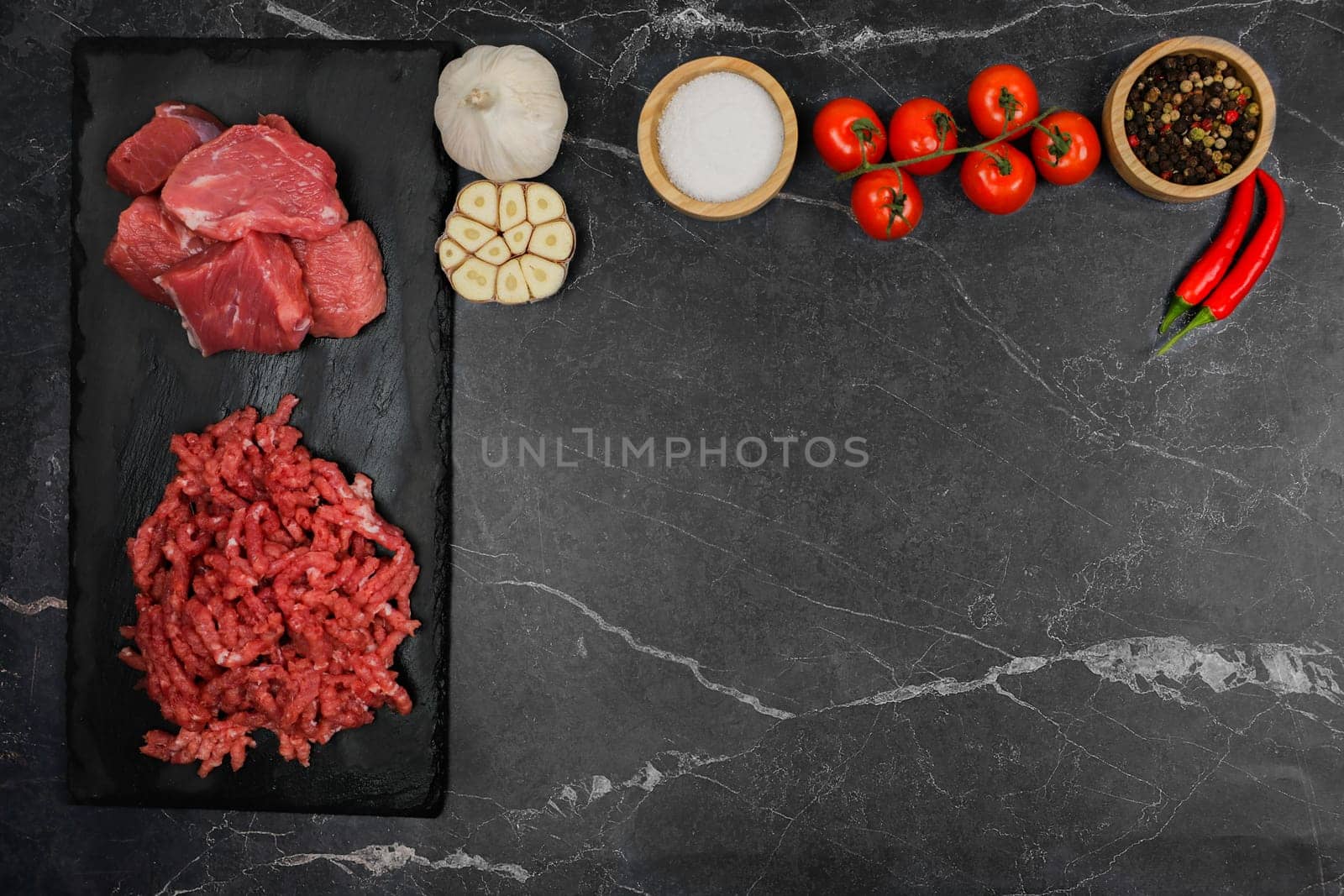 Top view of chunks of beef and minced meat lying on a black cutting board to the left, vegetables and condiments in wooden bowls on top with space for text. The concept of cooking, shredding meat, diet, natural products. High quality photo